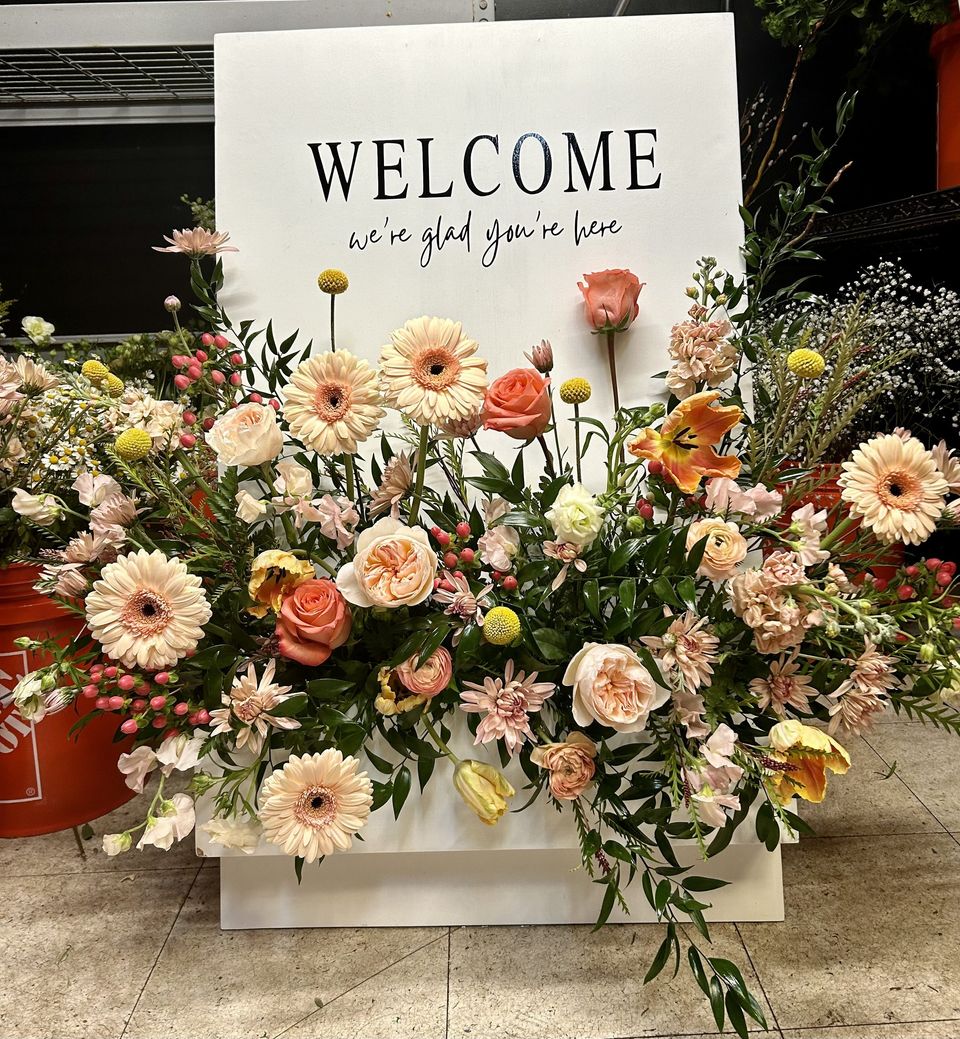 Welcome sign with a peach garden-style arrangement of gerberas, roses, and ranunculus