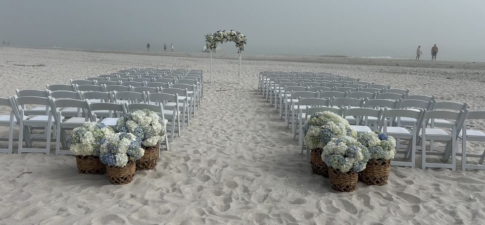 Beach ceremony with a white floral arch and hydrangea baskets along the aisle